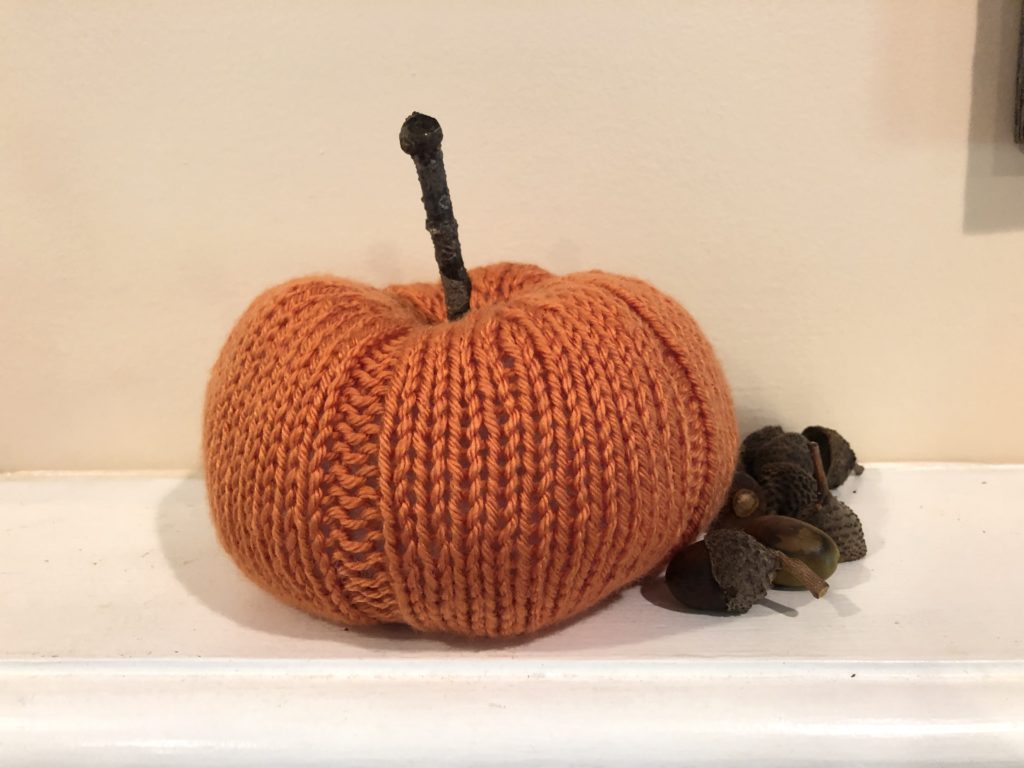 a knitted pumpkin and several acorns rest on a mantel