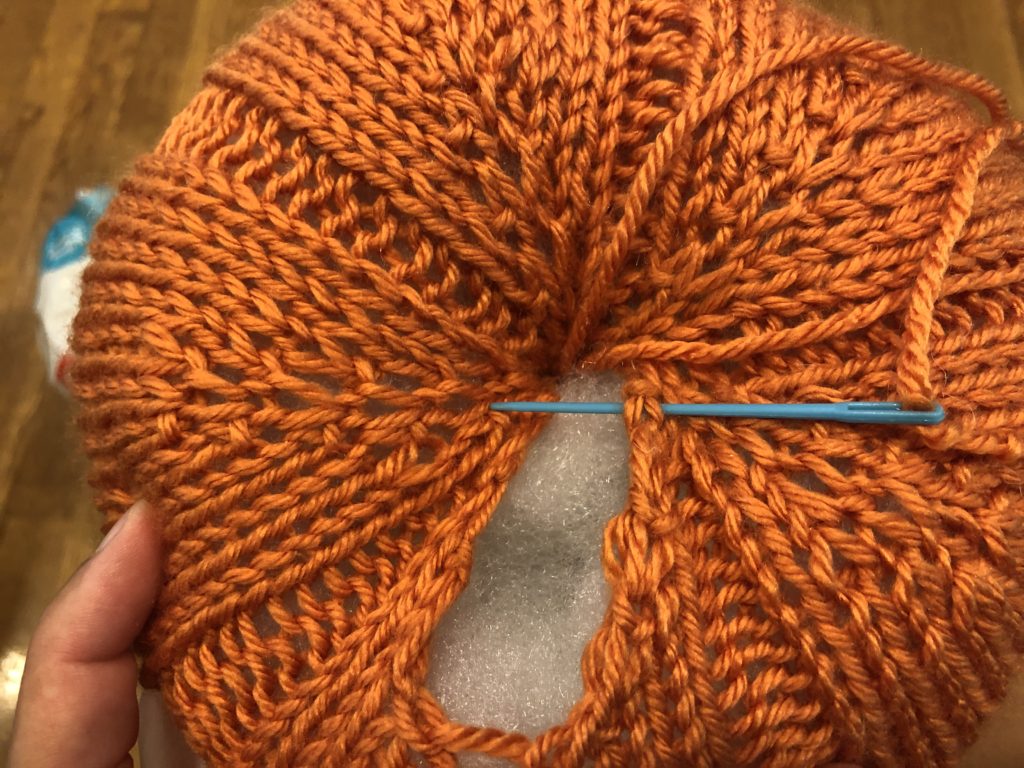 a knitted pumpkin full of stuffing with a needle in the process of sewing it closed