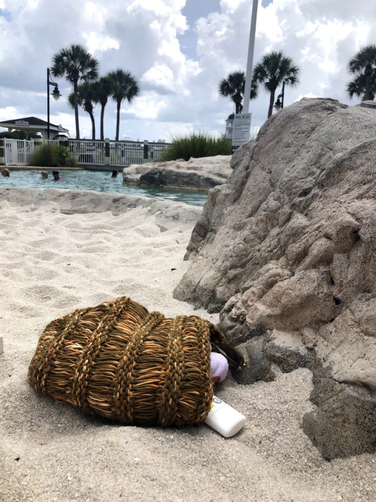 a brown beach bag is on its side in the sand, spilling sunscreen, sunglasses, and a towel