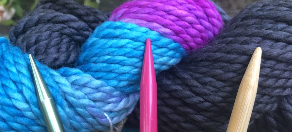 three knitting needles (aluminum, plastic, and bamboo) are displayed in front of a skein of yarn