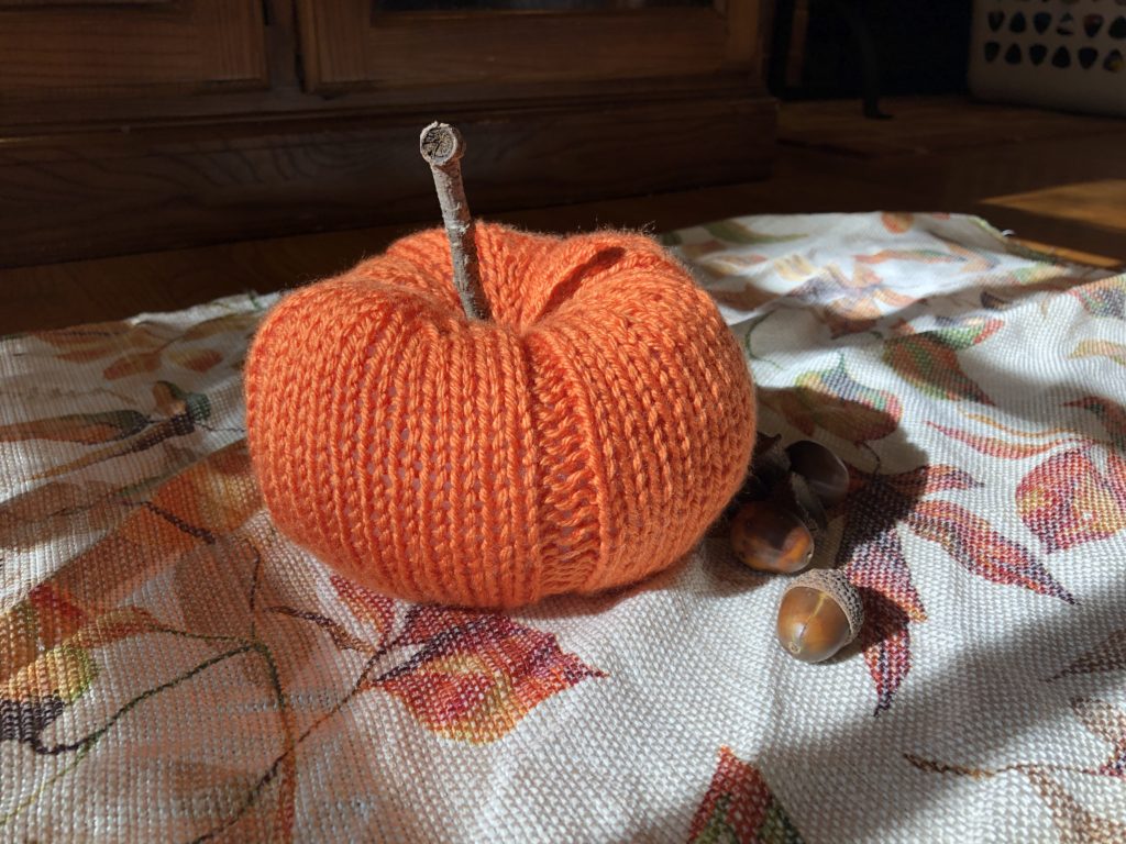 a knitted pumpkin with a stick stem rests next to an acorn on leaf-printed fabric