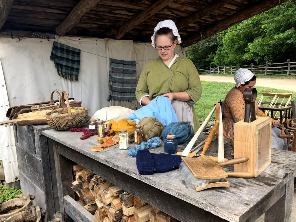 a historical interpreter sews behind a counter displaying wool, yarn, and natural dyes