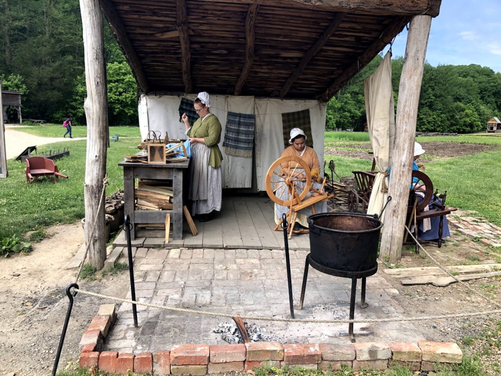 two historical interpreters sew and spin wool under a riverside shelter