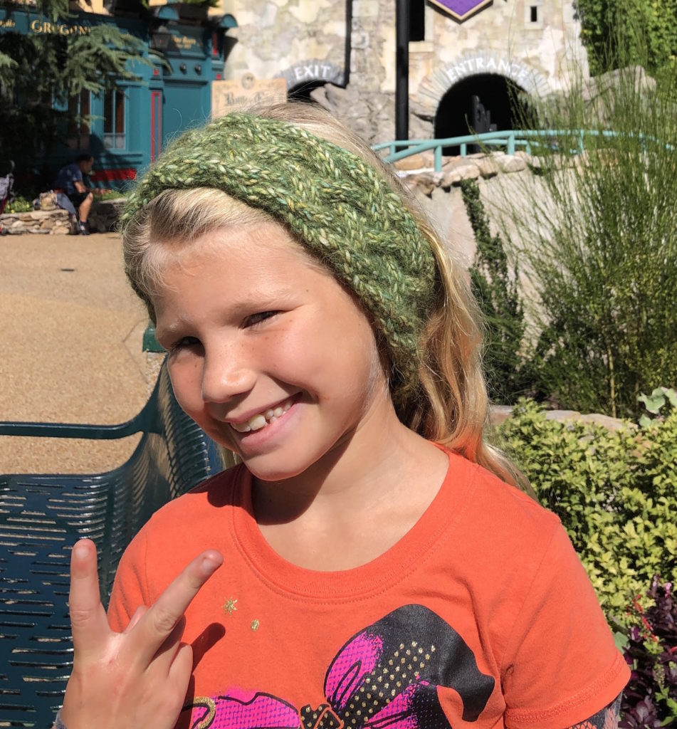 a young girl flashes the peace sign and smiles while wearing a hand knit cabled braided headband