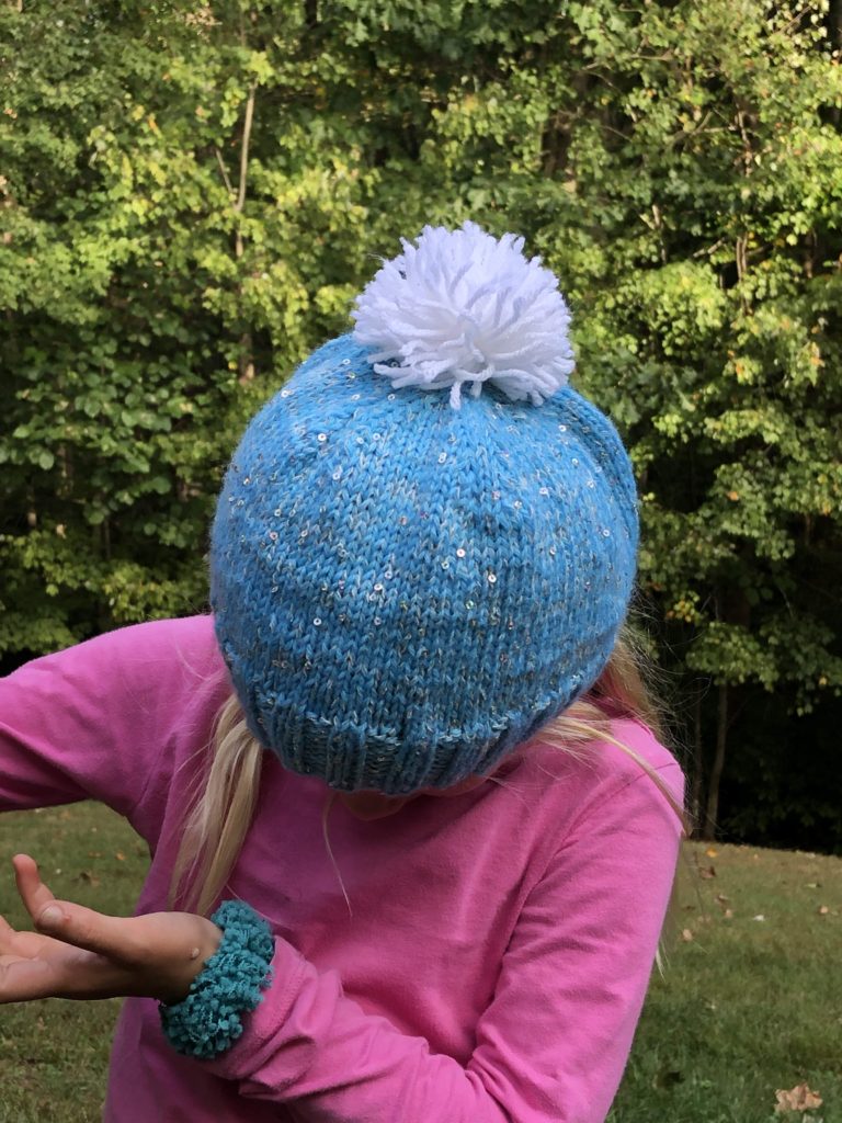 a child leans down to showcase the pompom on her knitted hat
