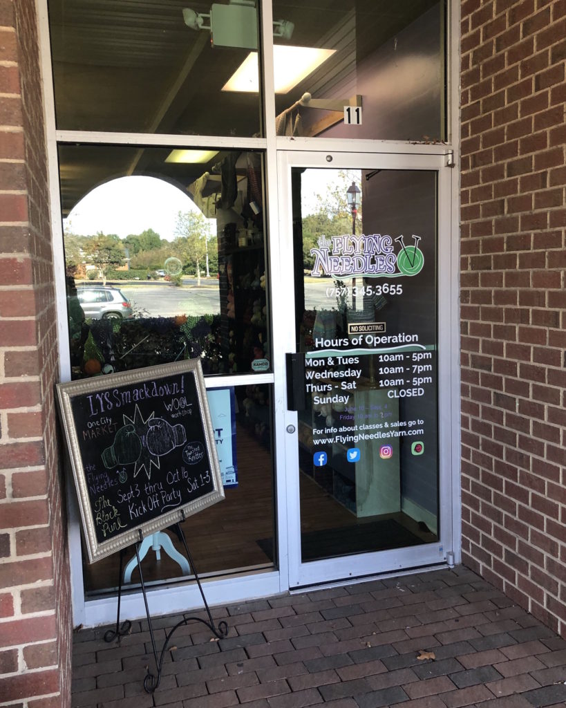 The Flying Needles yarn shop, a brick building with glass door and chalkboard display