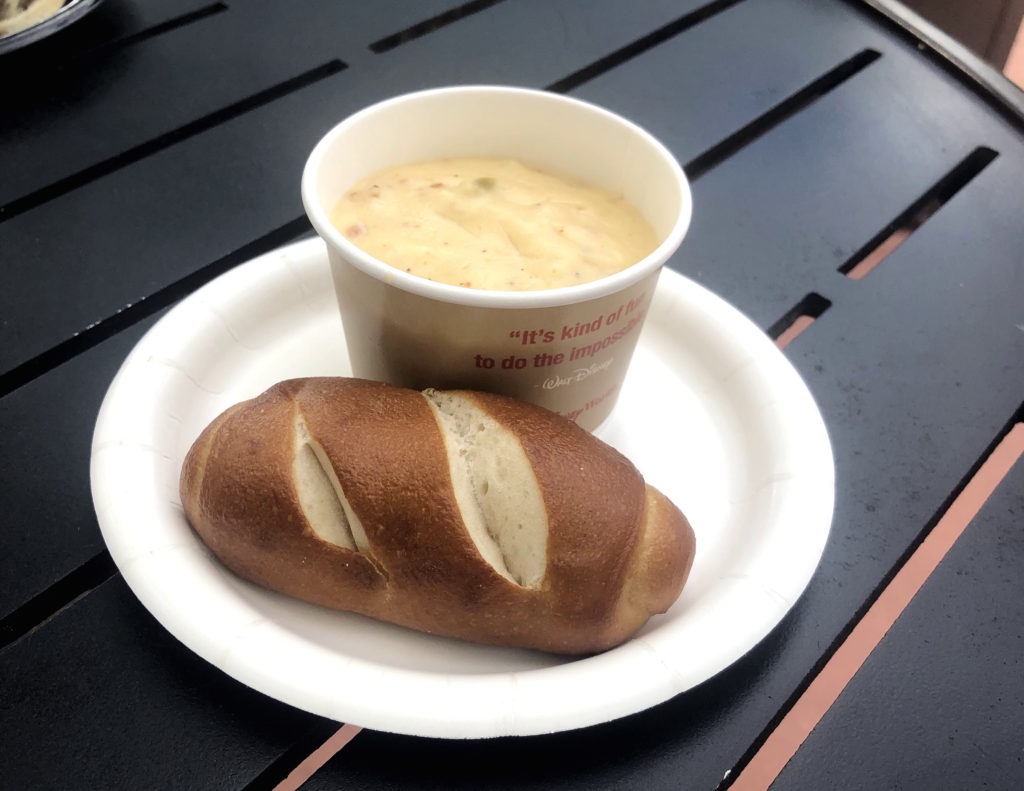 a bowl of Canadian cheddar cheese and bacon soup on a plate with a large pretzel roll