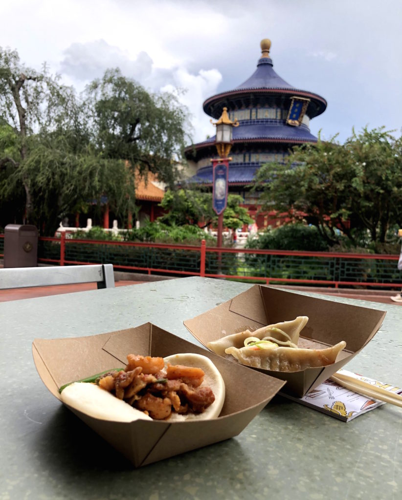 a close up of a Mala chicken and shrimp bao bun and dumplings with Chinese slaw on a table with the Temple of Heaven at Epcot's China Pavilion in the background