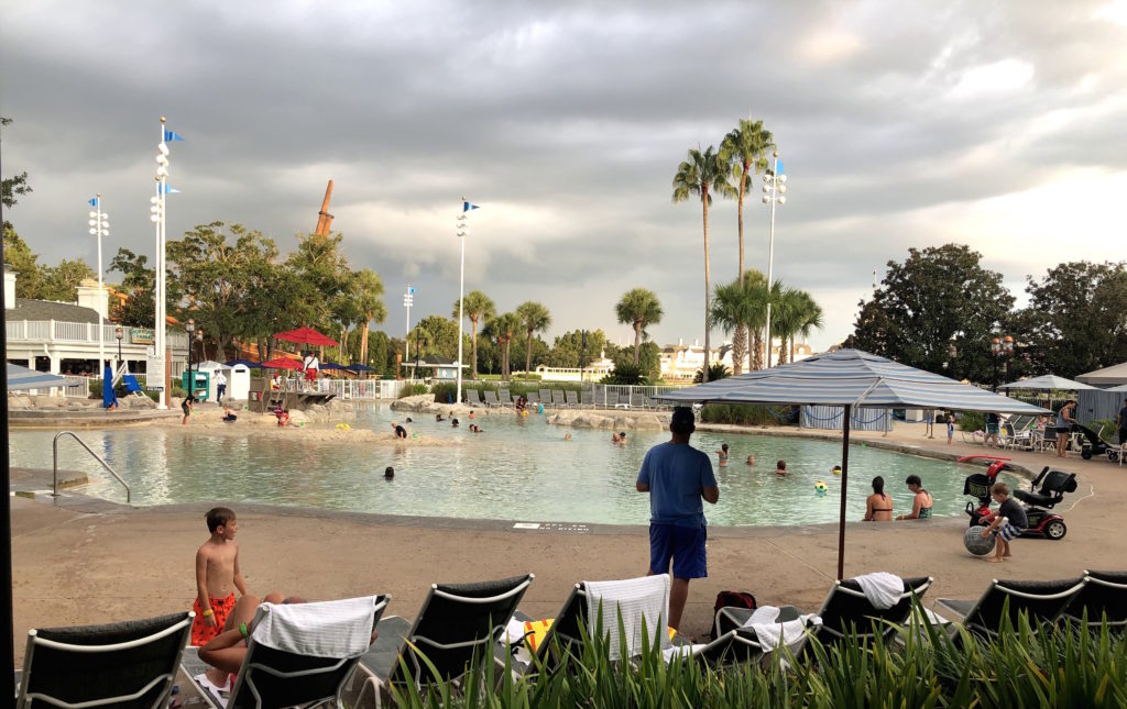 a large pool with a sand bottom surrounded by lounge chairs, palm trees, and people