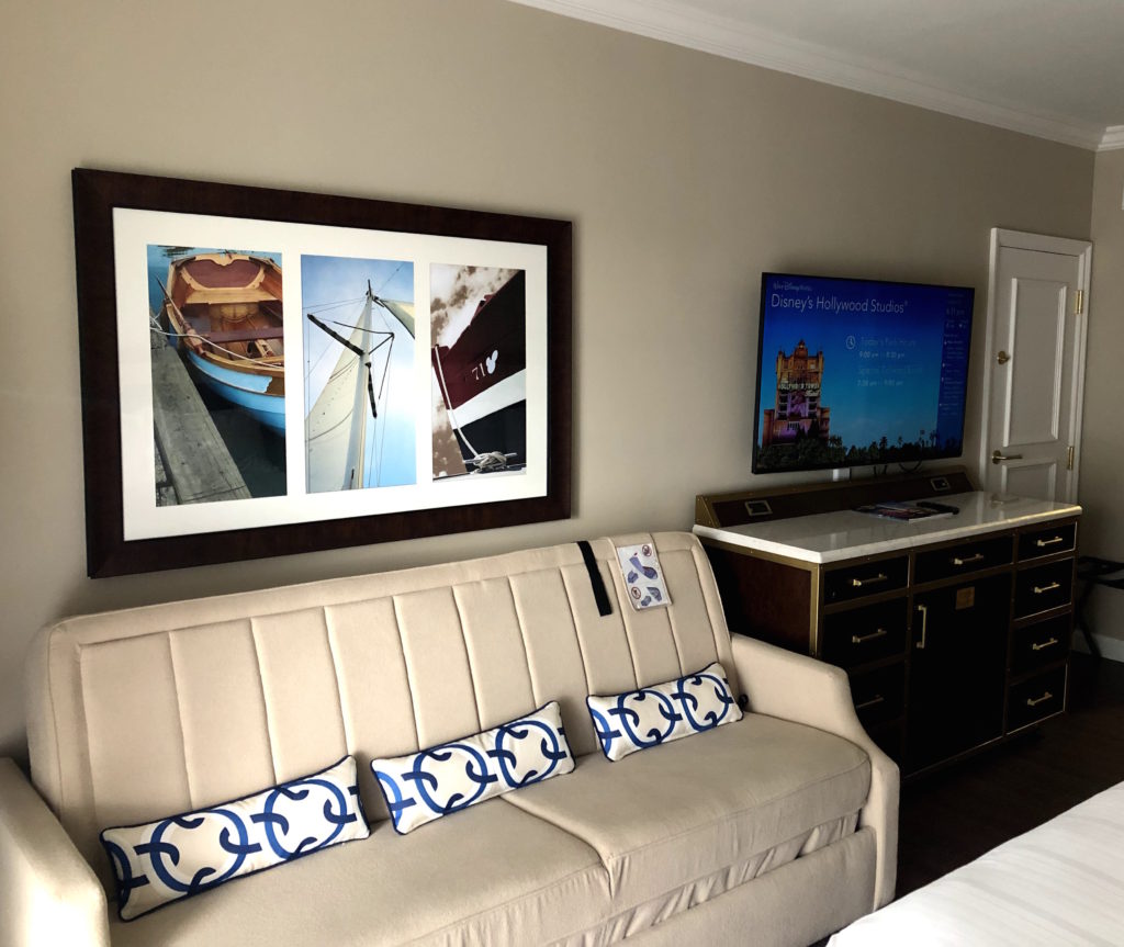 a hotel room featuring a sofa with throw pillows, a television over a dresser, and nautical art