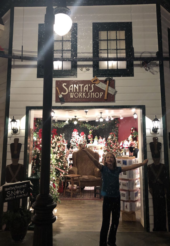a child standing with her arms up in snowfall under a sign that reads Santa's Workshop