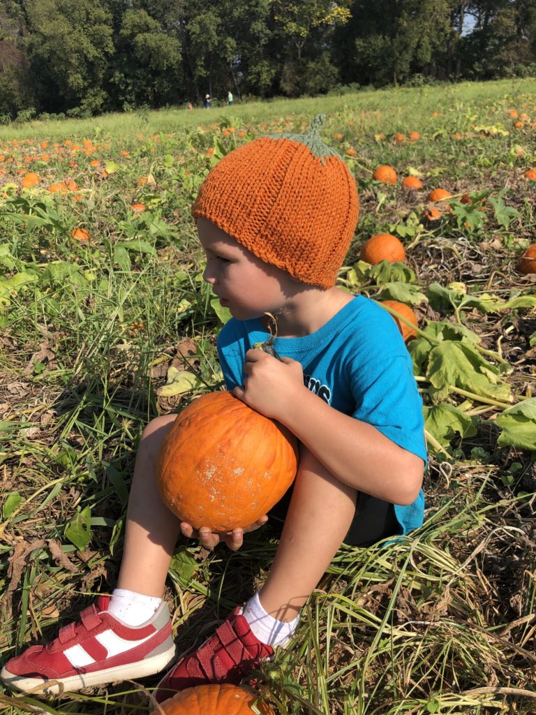 a young boy wearing a knitted pumpkin hat sits in a pumpkin patch while holding a pumpkin