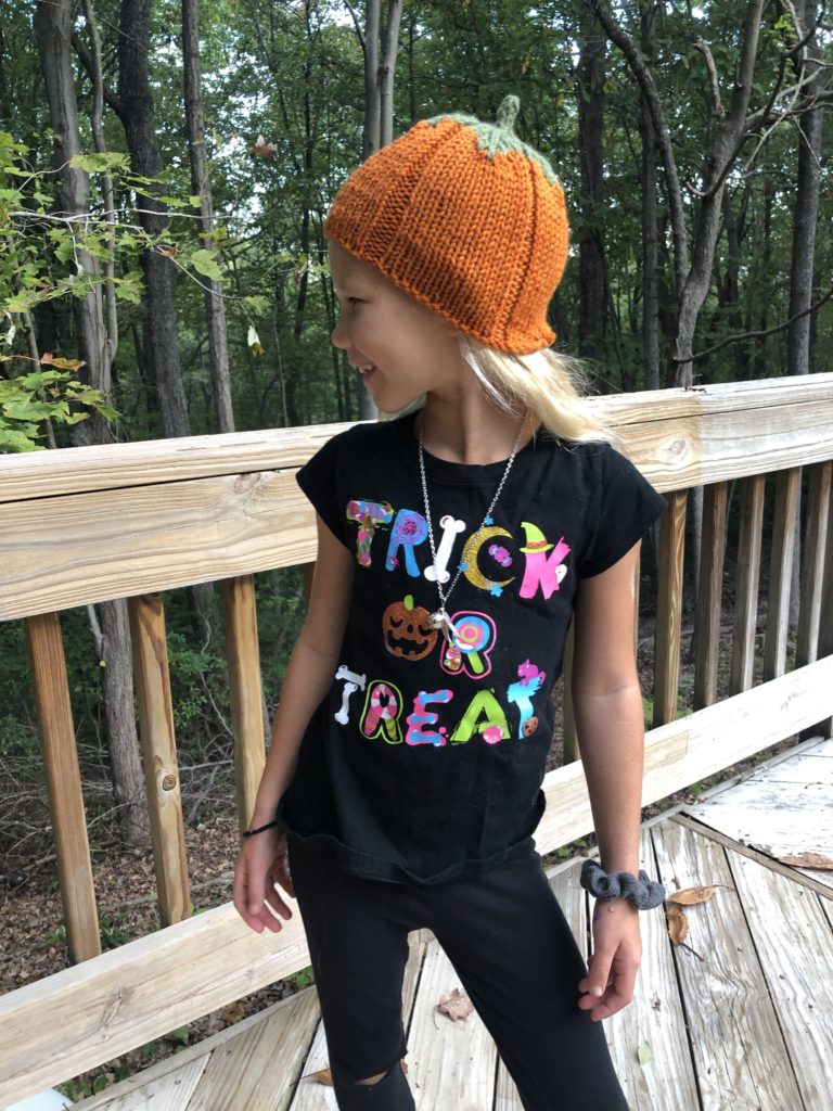 a young girl in the woods wears a knitted pumpkin hat with a shirt that says "Trick or Treat"