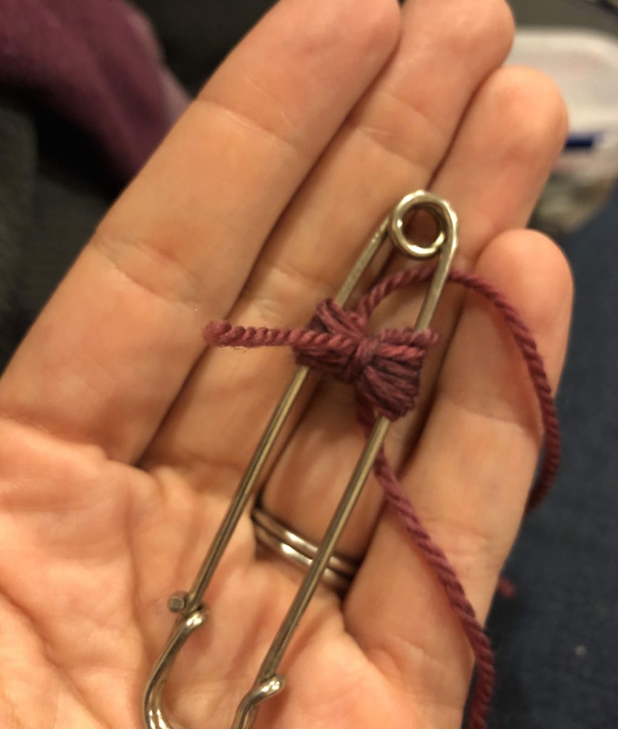 a hand holding a large safety pin with yarn wrapped around it and tied through the center