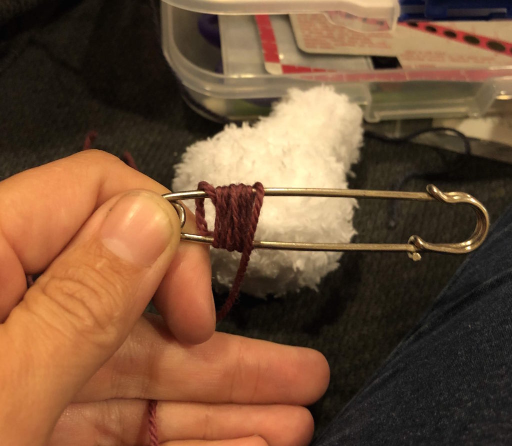 a hand holding a large safety pin, demonstrating how to wrap it with yarn for snowman knitting pattern