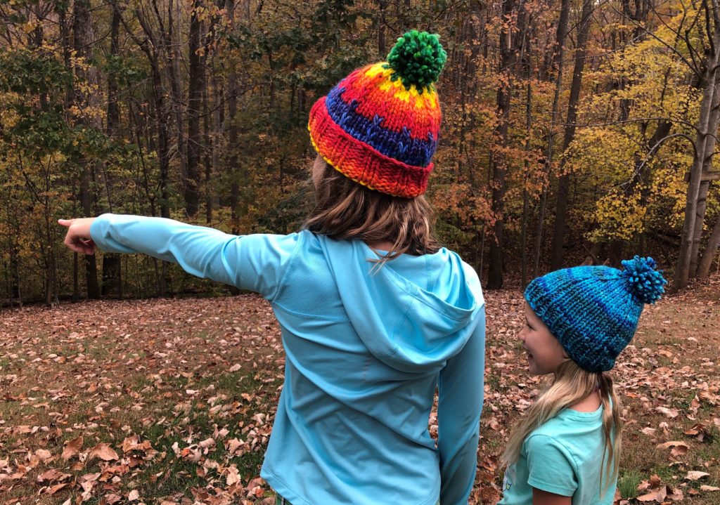 a child wearing a rainbow knitted hat with a green pompom points to something in the distance while a child in a blue knitted hat looks on