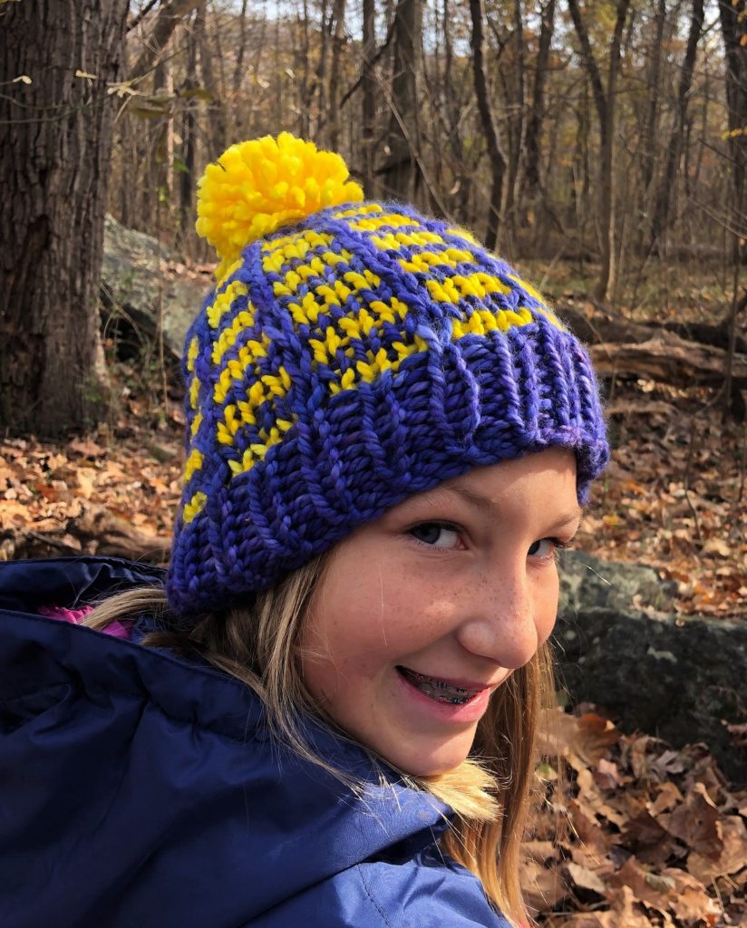 a young girl wears a purple and yellow brick style striped hat