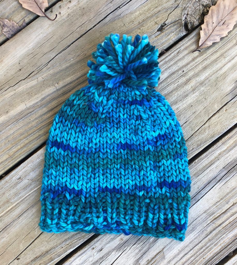 a vibrant multi-color blue knitted hat with pompom on a wood panel background