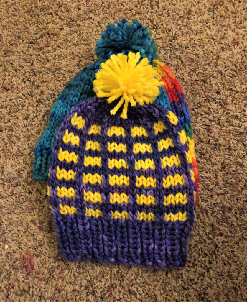 a purple and yellow brick stripe style knitted hat rests on top of two other multicolored hats