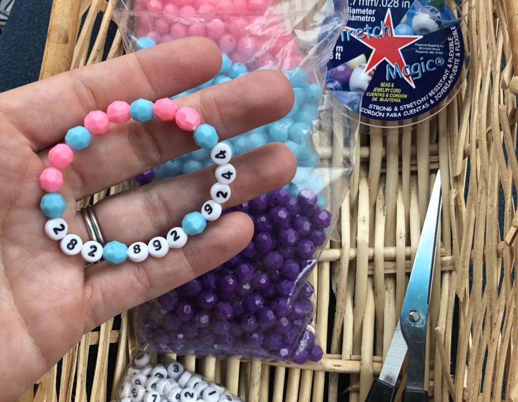 a child's hand holds a bracelet with beads that read 282 892 9244 with beads, scissors, and stretch magic elastic in the background