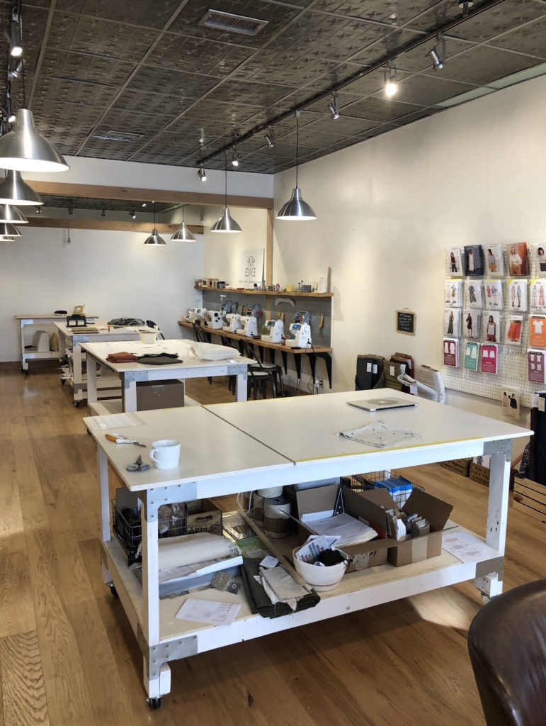 The Maker's Lounge: large multi-use sewing tables, a row of sewing machines, and a wall lined with sewing patterns