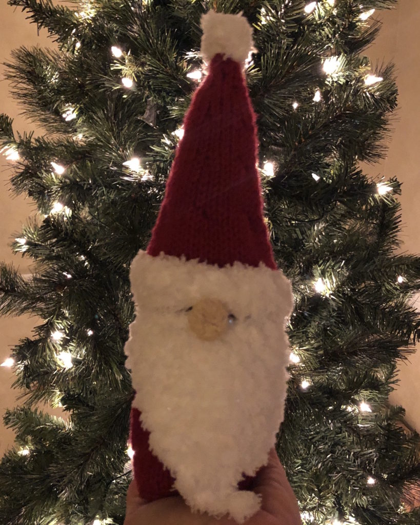 a Santa Claus bottle cozy in front of a lit Christmas tree