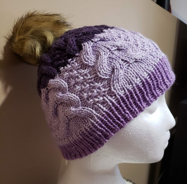 a purple hat with cable details and a faux fur pompom adorns a Styrofoam head