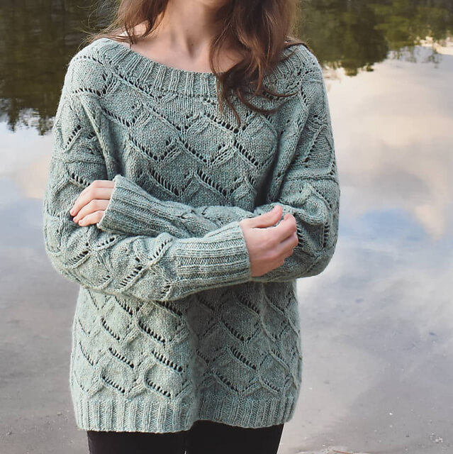 a woman stands in front of a lake with her arms crossed wearing the waterscapes knitted pullover