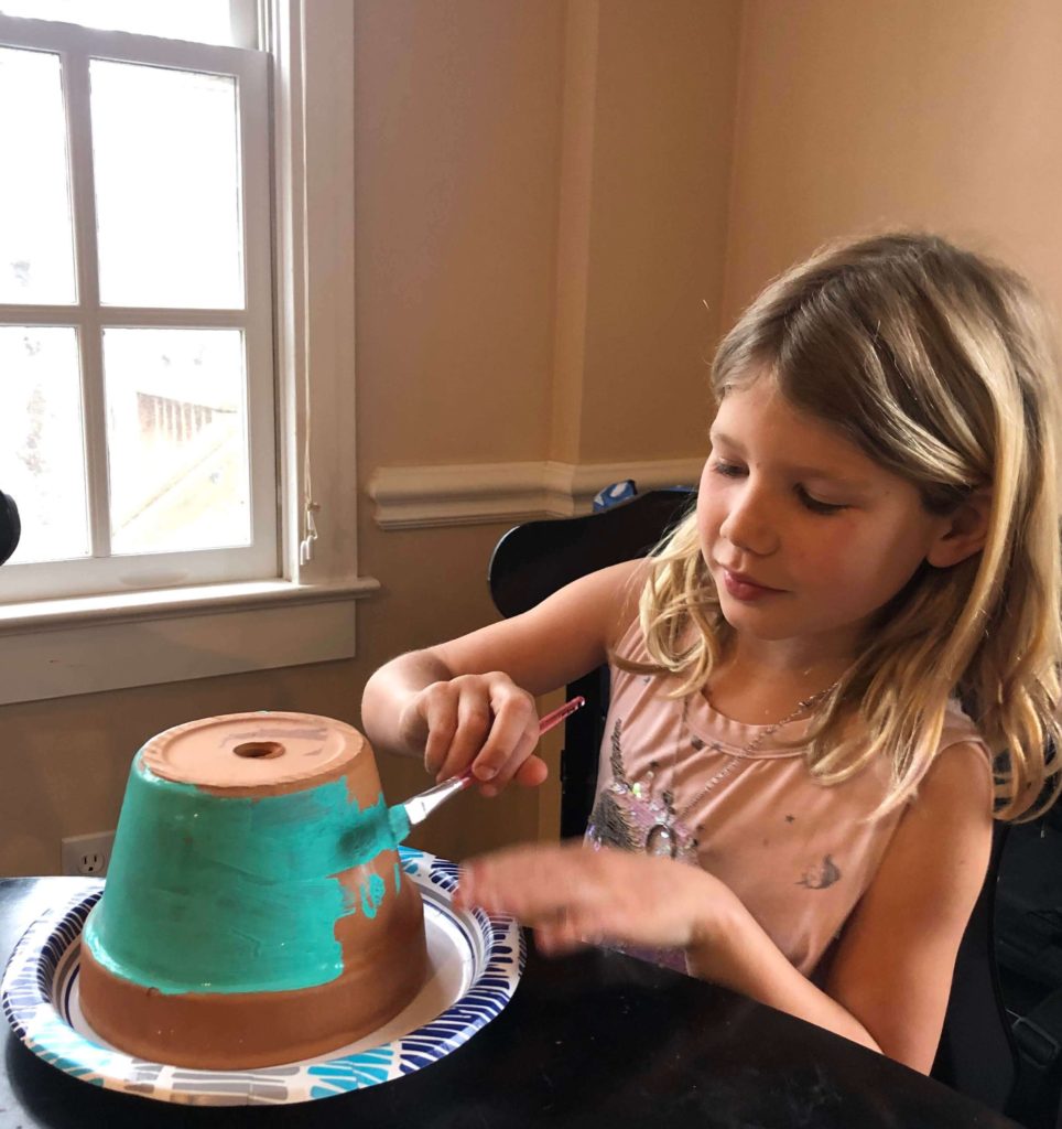 a young girl carefully paints an upside-down terracotta flowerpot on a paper plate