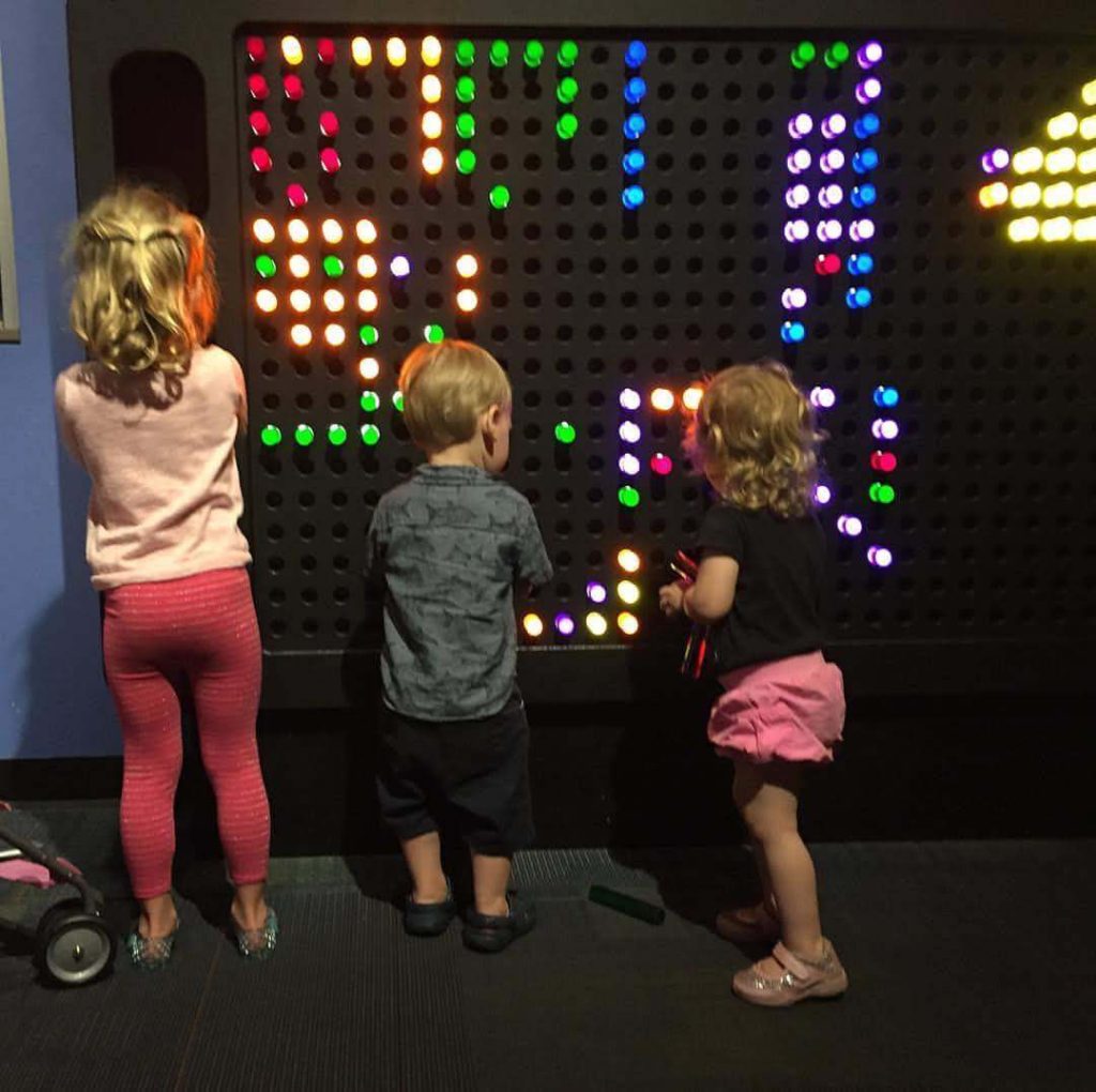 three children take turns swapping multicolored pegs in a giant Lite Brite