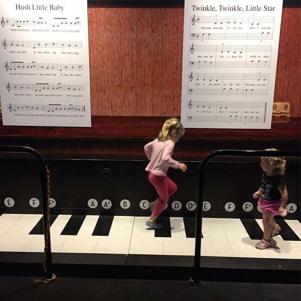two children dance on a giant keyboard in front of sheets of music larger than they are