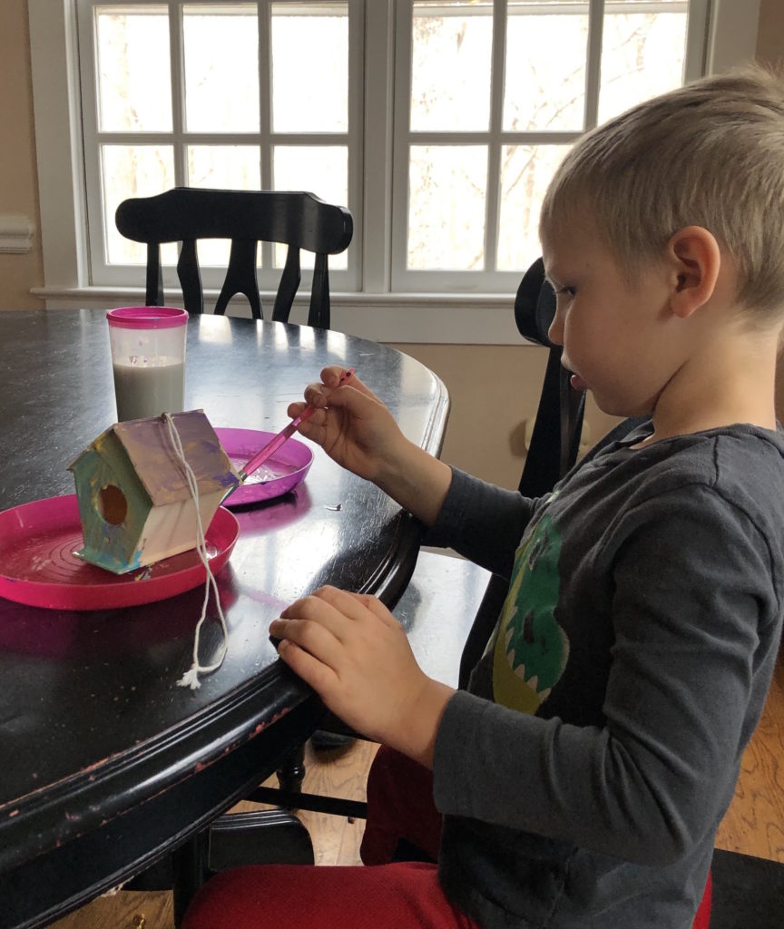 a young boy carefully paints a wooden birdhouse at his dining room table