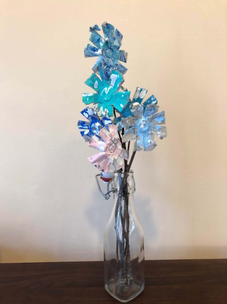painted flowers made of egg cartons and sticks rest in a glass bottle as a vase