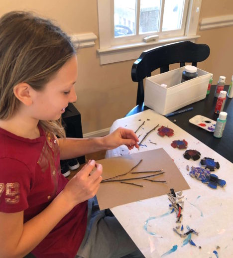 a young girl glues sticks to a cardboard background and prepares to position her egg carton flowers