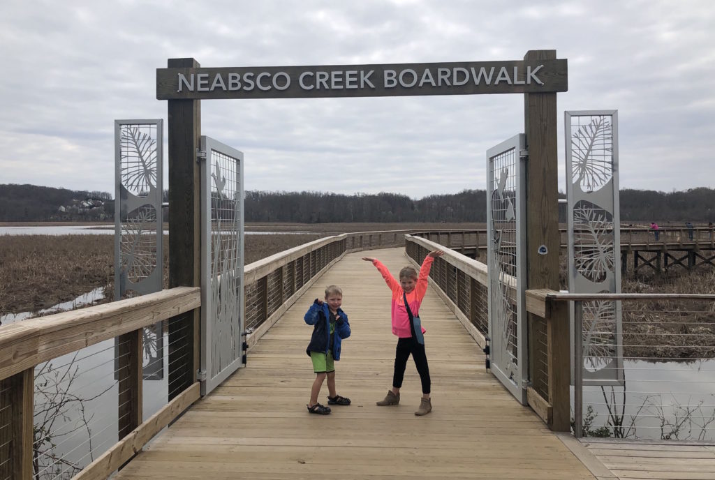 two children pose gleefully with outstretched arms on a wooden bridge underneath a sign that reads "Neabsco Creek Boardwalk"