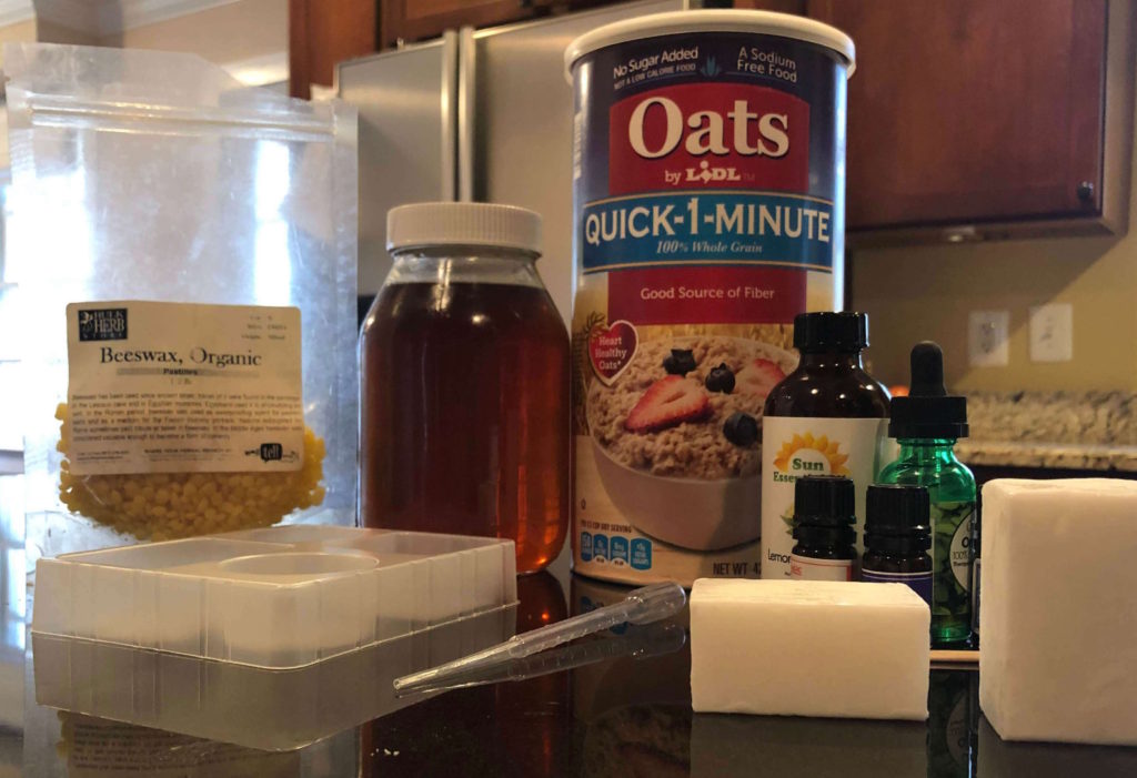 soap ingredients and soap-making supplies, including a mold, beeswax, honey, oats, essential oils, and pipette, are piled on a kitchen counter