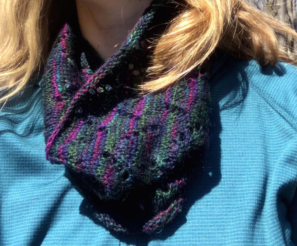 a woman wears a a multicolored woolen cowl with v-shaped eyelets