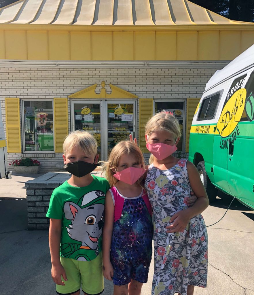 three children in cloth masks pose in front of a small, brick building covered in lemons