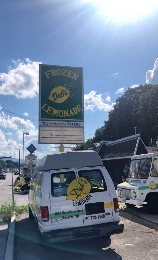The sun shines down on a large sign that reads "Deb's Frozen Lemonade - Yes - We're Open - Snacks" while two mobile lemonade trucks charge their batteries
