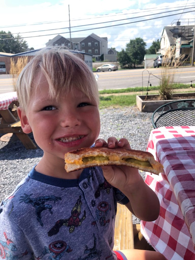 a young, smiling boy holds a grilled cheese sandwich with pickles and smiles