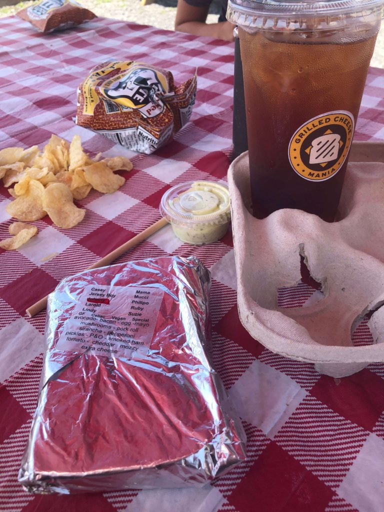 a checkered tablecloth with sandwiches, chips, and iced tea bearing the Grilled Cheese Mania logo