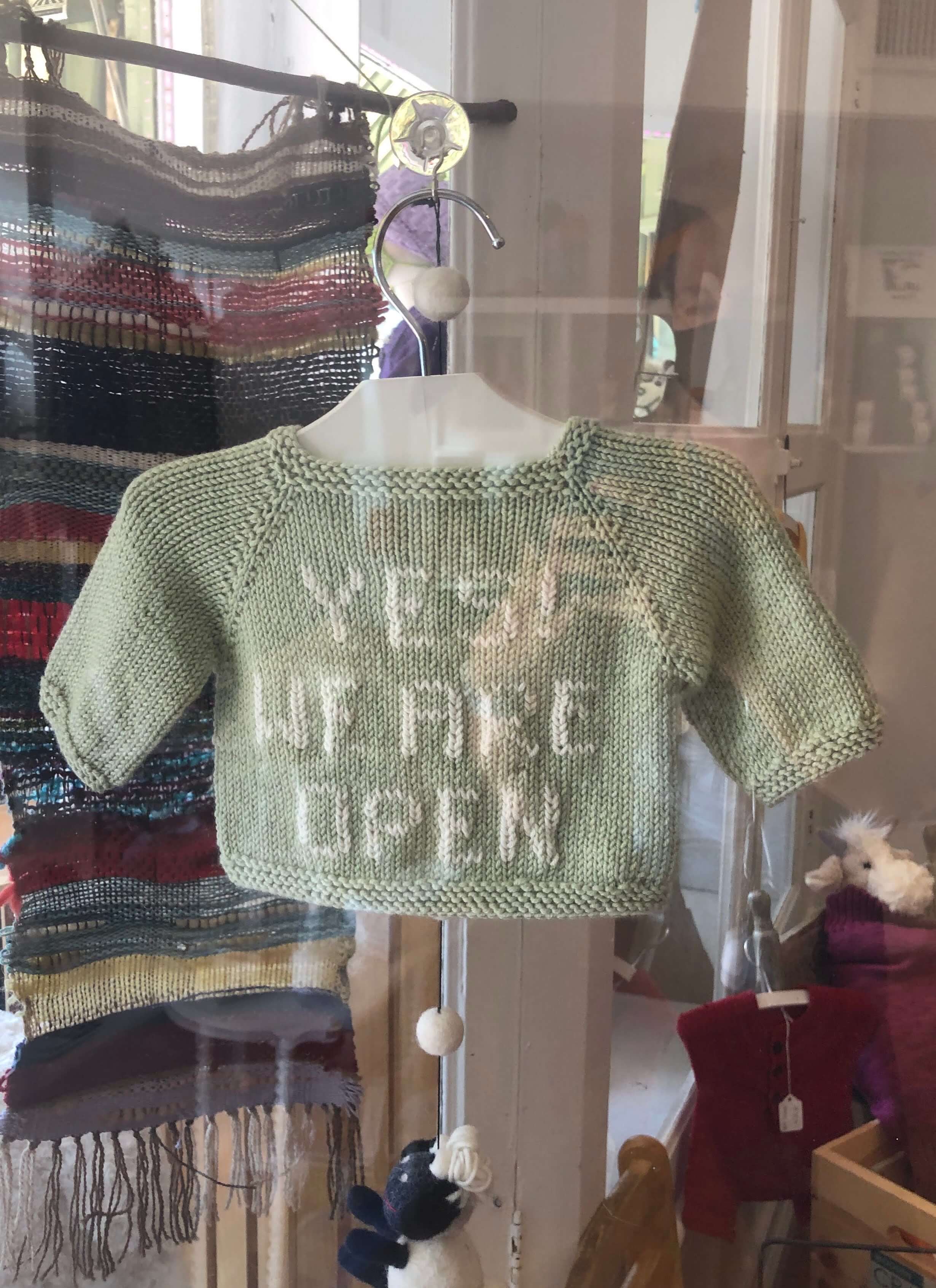 a knitted sweater photographed through the shop front glass reads "YES WE ARE OPEN"