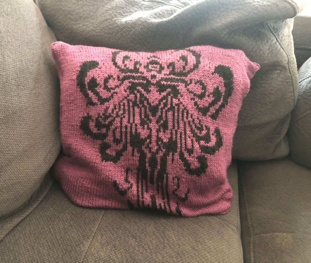 a knitted pillow with a creepy-looking face stitched into it rests on a couch