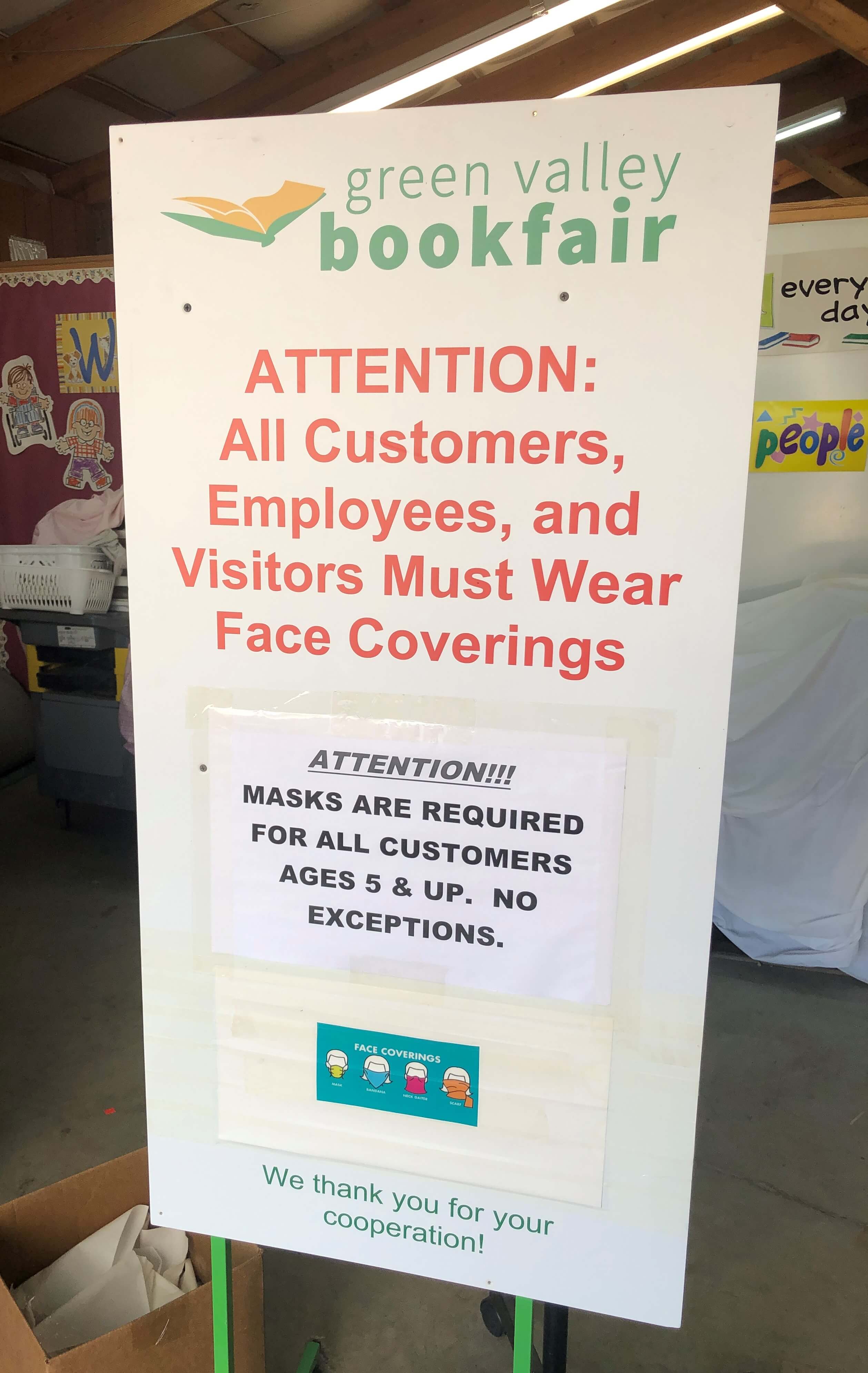 a sign that reads "Green Valley Book Fair Attention: All Customers, Employees, and Visitors Must Wear Face Coverings Attention!!! Masks are required for all customers ages 5 & up. No exceptions. We thank you for your cooperation!"