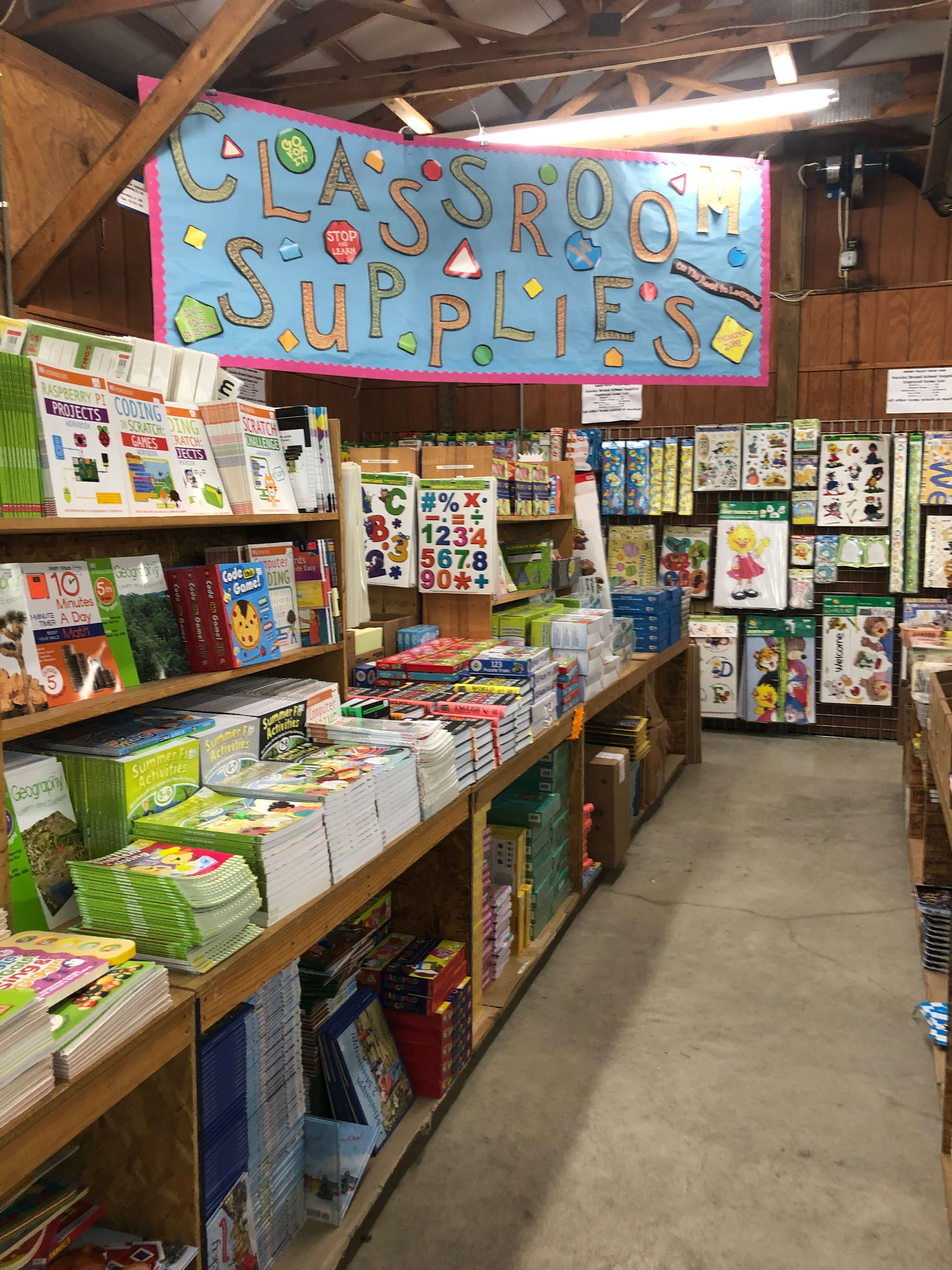 Books, manipulatives, stickers, and bulletin board accessories line the shelves. A large banner overhead reads CLASSROOM SUPPLIES.