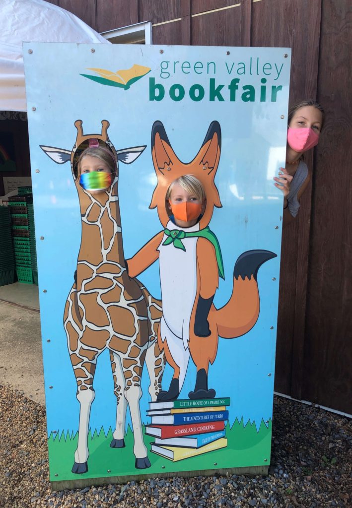 three children peek around a Green Valley Book Fair face-in-hole cutout featuring a giraffe and a fox