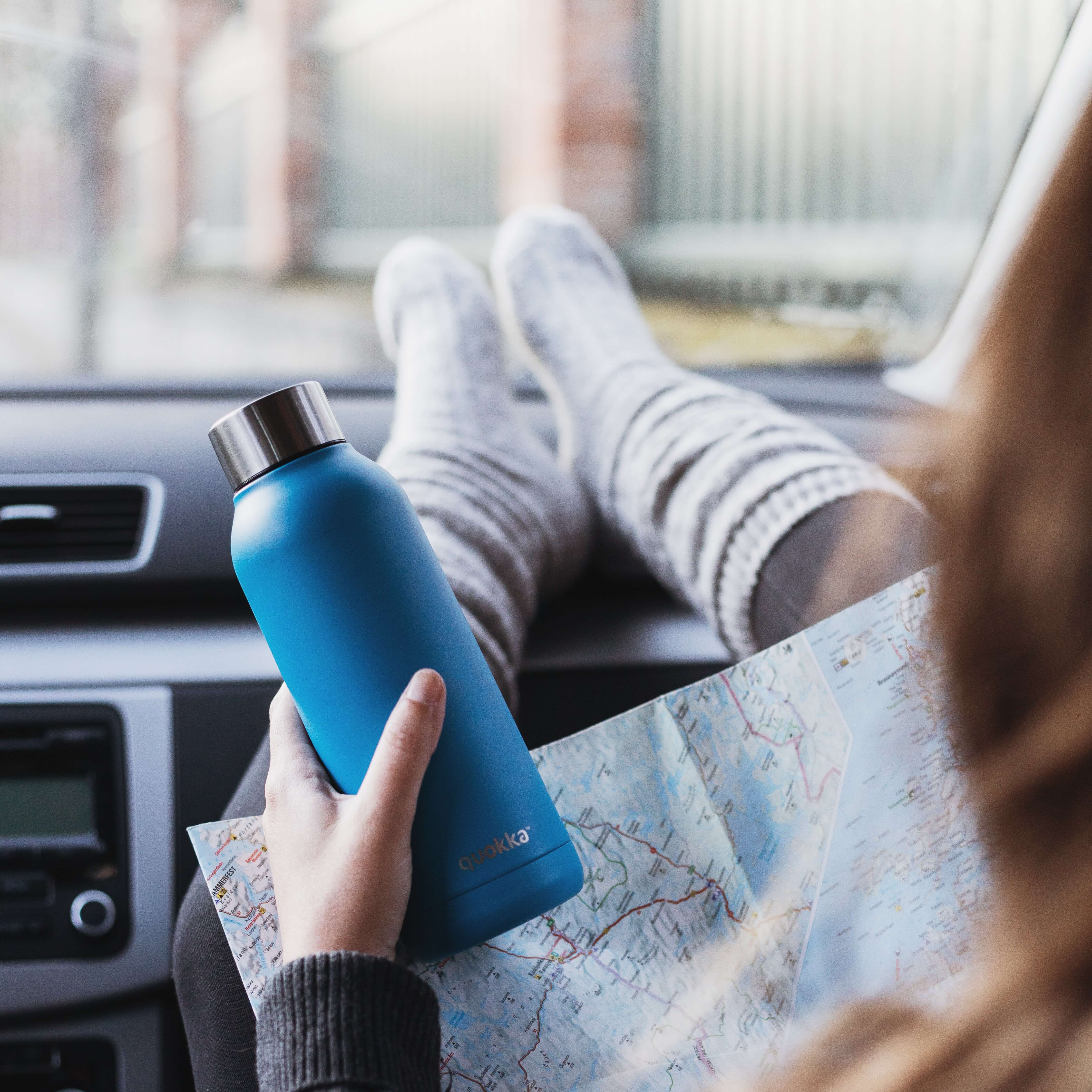 a woman consults a map while holding a water bottle and resting her feet on the dashboard of the car