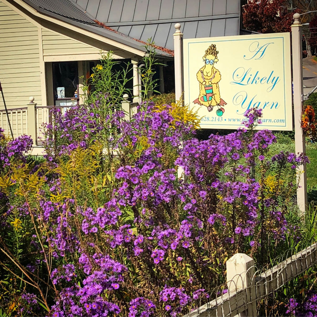 The exterior of a cottage as seen across the flower garden. A sign reads "A Likely Yarn"