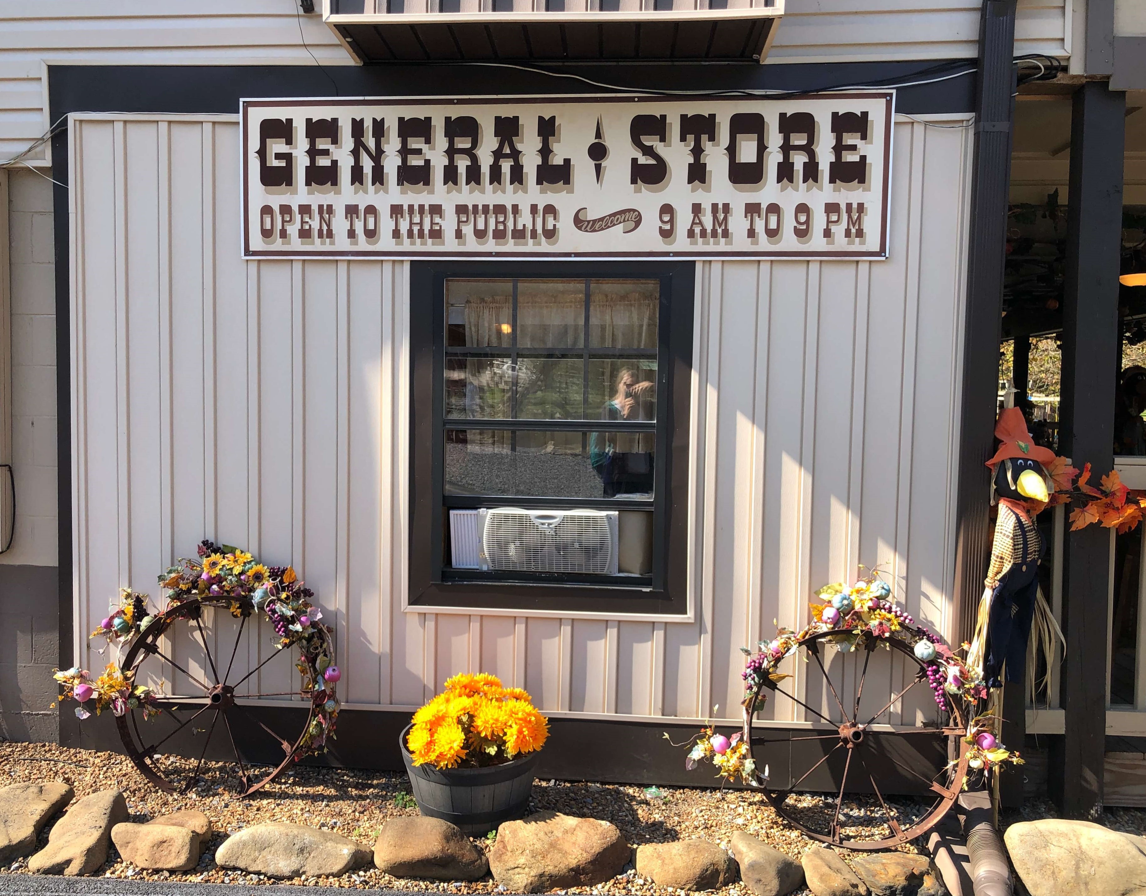 outside a shop with flower-decorated wagon wheels, a sign reads GENERAL STORE, OPEN TO THE PUBLIC, 9 AM to 9 PM