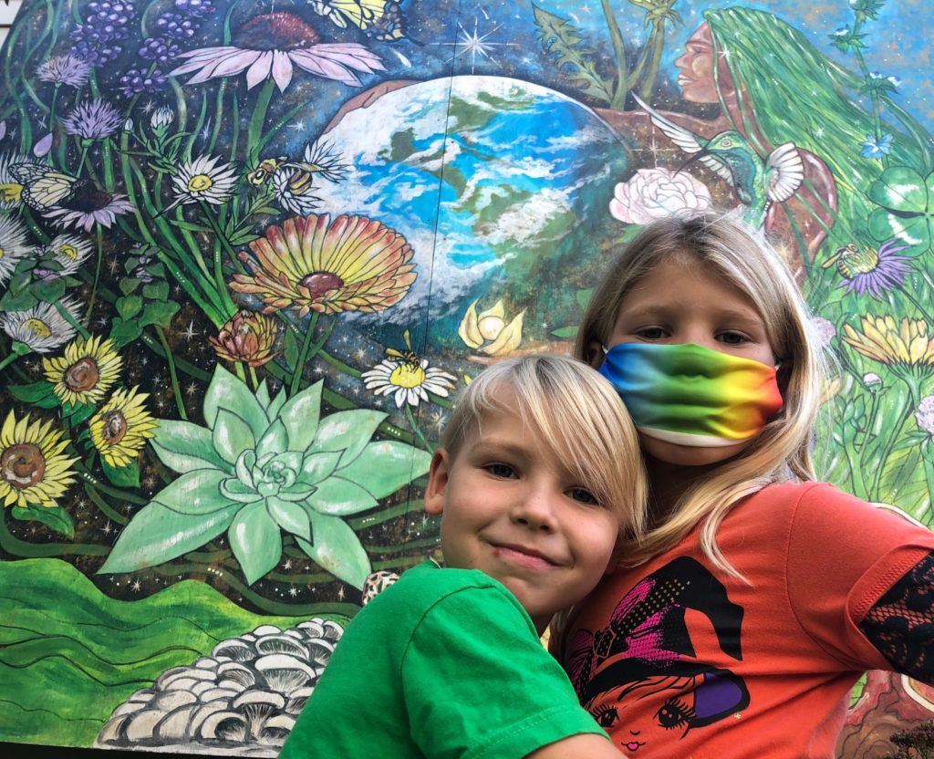 two children smile in front of a colorful mural of Gaia, earth, and a garden